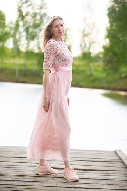 A beautiful elderly woman in a flowing pink dress stands on a wooden bridge near the river