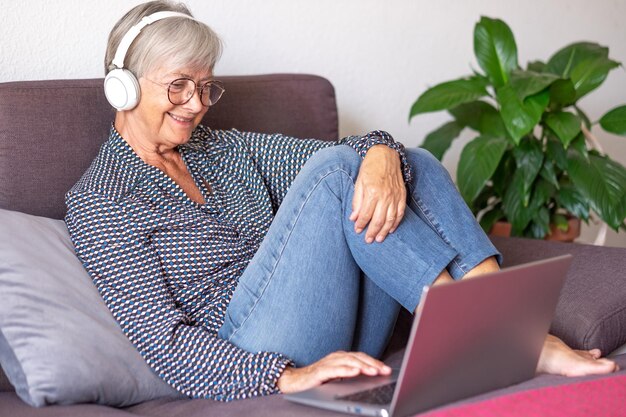 Beautiful elderly woman using laptop at home Sitting on sofa in living room wearing headphones Senior lady enjoying tech and social