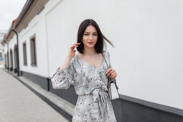 Beautiful elegant fashionable woman model with a bob haircut in a stylish summer floral dress with a bag walking in the city
