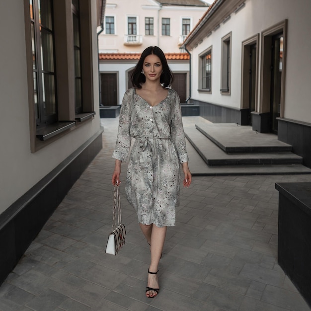 Beautiful elegant fashionable woman in a stylish vintage dress with a floral pattern with a bag and heels walking in the city