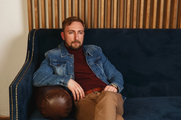 Beautiful elegant fashionable young man in a blue jacket sits on sofa