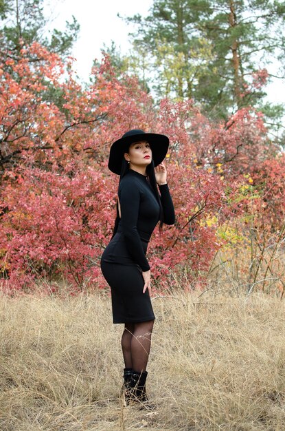 Beautiful elegant woman in a black hat standing fulllength in a park in autumn