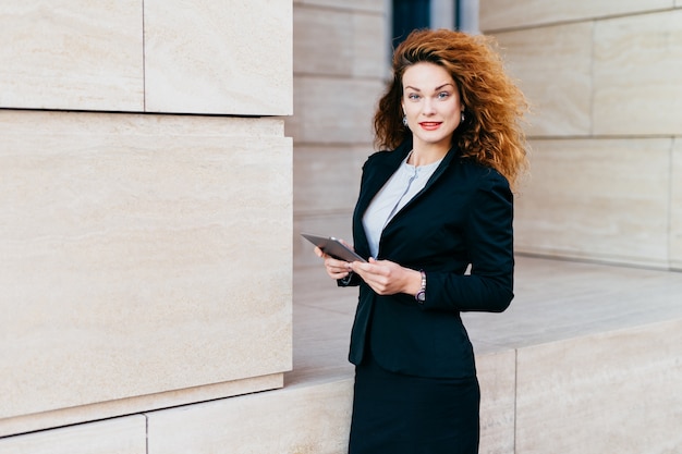 Beautiful elegant young businesswoman in black costume, having bushy curly hair and appealing appearance