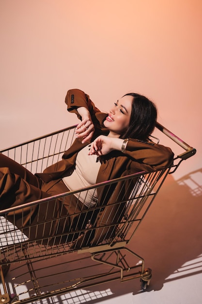 Beautiful elegant young woman model in nice stylish brown suit posing in shopping cart on gray background Young elegant woman in a suit