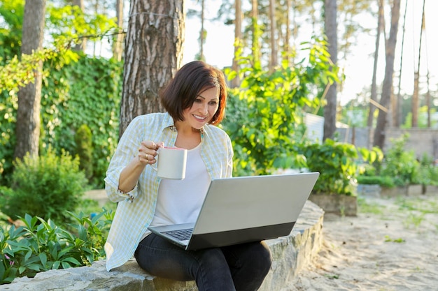 Beautiful emotional middle aged woman relaxing in garden with cup of tea and laptop