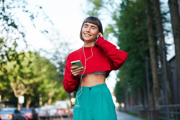 Beautiful emotional woman listening music on the street, cute millenial woman in red stylish sweater using erphones on the modern