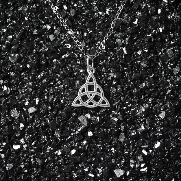 Beautiful ethnic Scandinavian Celtic Claddagh Triquetra Silver jewelry Necklace, charmed symbol of magic on black crashed glass background.