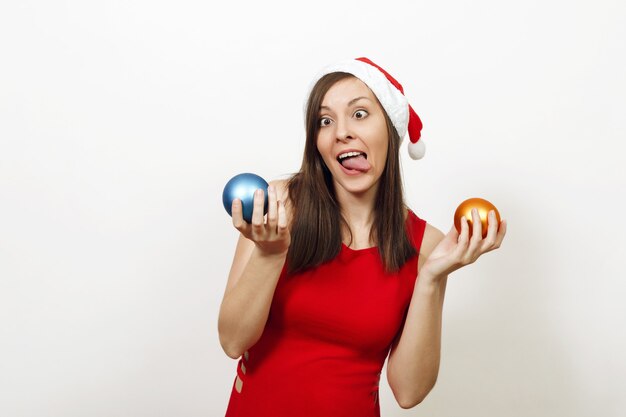 Beautiful European funny happy woman with healthy skin and charming smile in red dress and Christmas hat holding two tree toys on white background. Santa girl isolated. New Year holiday 2018 concept.