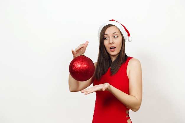赤いドレスと白い背景の上の大きな光沢のある木のおもちゃを保持しているクリスマスの帽子の魅力的な笑顔で美しいヨーロッパの驚きの若い幸せな女性。孤立したサンタの女の子。 2018年の年末年始のコンセプト