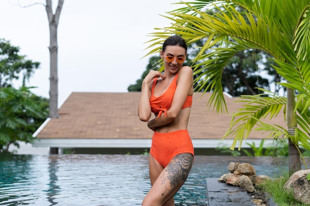 Beautiful european women in two pieces orange bikini swimsuit in perfect shape good body tanned with trendy sunglasses outside at tropical swimming pool and palm leaf tree