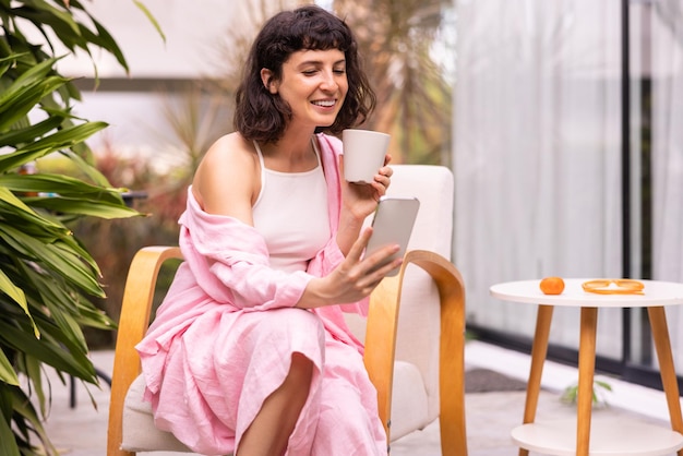 Beautiful european young brunette girl reads morning news through smartphone drinking coffee sitting on terrace Relaxed lifestyle concept