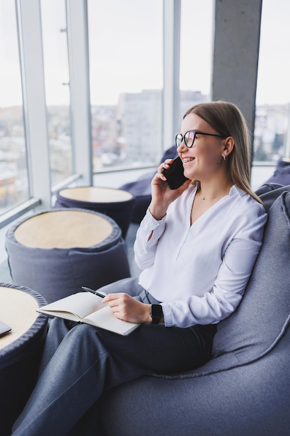 A beautiful European young business woman in a coffee shop sits with a mobile phone and makes notes in a notebook Wearing an elegant casual suit and sitting with a laptop in a public cafe