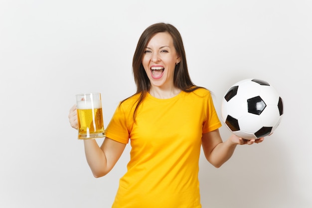 Beautiful European young cheerful woman, football fan or player in yellow uniform holding pint mug of beer, soccer ball isolated on white background. Sport, play football, healthy lifestyle concept.