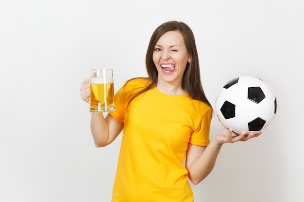 Beautiful European young cheerful woman, football fan or player in yellow uniform holding pint mug of beer, soccer ball isolated on white background. Sport, play football, healthy lifestyle concept.
