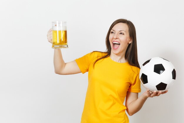 Beautiful European young cheerful woman, football fan or player in yellow uniform holding pint mug of beer, soccer ball isolated on white background. Sport, play football, healthy lifestyle concept.