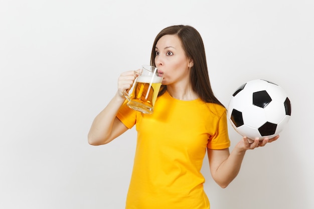 Beautiful European young cheerful woman, football fan or player in yellow uniform holding pint mug of beer, soccer ball isolated on white background. Sport, play football, healthy lifestyle concept.