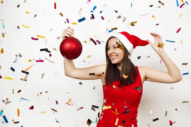 Beautiful European young happy woman with charming smile in red dress and Christmas hat holding big tree toy on white background and confetti. Santa girl isolated. New Year holiday 2018 concept.