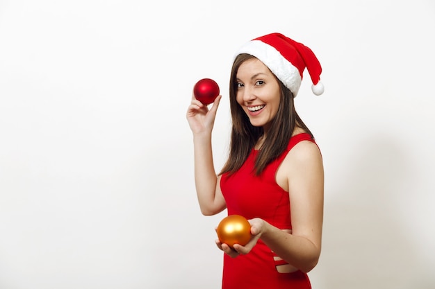 Beautiful European young happy woman with healthy skin and charming smile in red dress and Christmas hat holding two tree toys on white background. Santa girl isolated. New Year holiday 2018 concept.