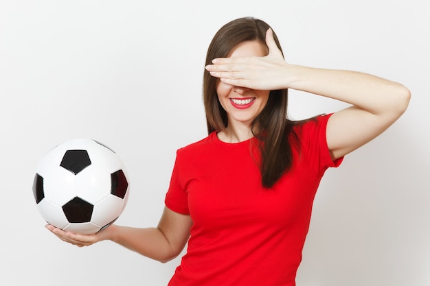 Beautiful european young woman, football fan or player in red\
uniform hold classic soccer ball, cover eyes by hand isolated on\
white background. sport play football, health, healthy lifestyle\
concept.