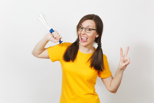 Beautiful European young woman with two fun pony tails, fan or player in glasses, yellow uniform show victory sign, hold football pipe isolated on white background. Sport, healthy lifestyle concept.