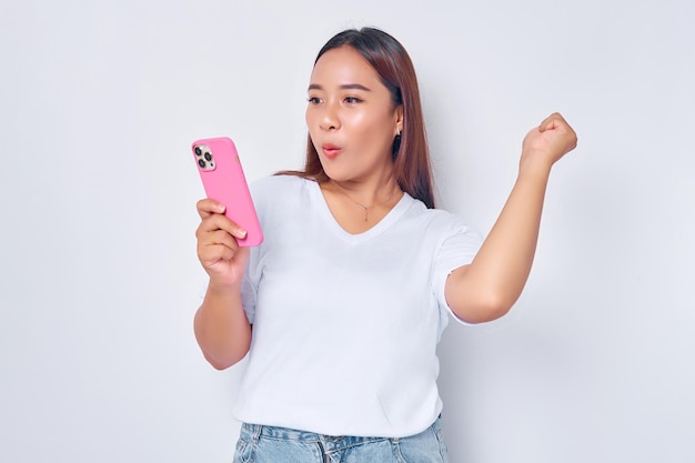 Beautiful excited Asian girl wearing casual white tshirt holding mobile phone raising her fist celebrating good luck isolated on white background People lifestyle concept