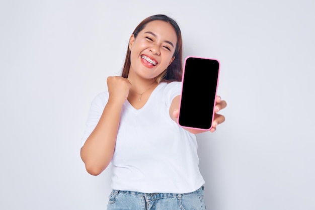 Beautiful excited Asian girl wearing casual white tshirt showing mobile phone with blank white screen raising fist celebrating good luck isolated on white background People lifestyle concept