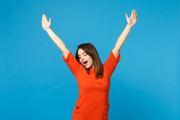 Beautiful excited brunette young woman wearing red orange dress raised hands up, winner say Yes isolated over blue wall background studio portrait. People lifestyle fashion concept. Mock up copy space