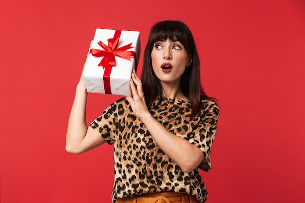 Beautiful excited shocked young woman dressed in animal printed shirt posing isolated over red wall holding present box.