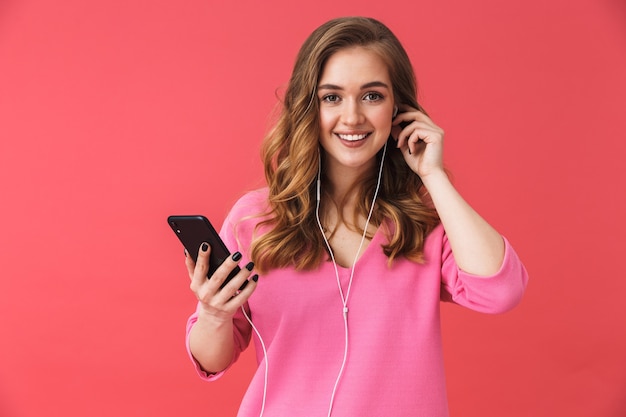 Фото Красивая возбужденная молодая девушка в повседневной одежде стоит изолированно над розовой стеной, слушает музыку в наушниках и держит мобильный телефон