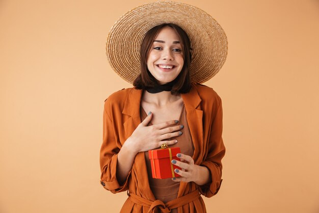 밀짚 모자와 여름 옷을 입고 베이지색 벽 위에 격리된 채 선물 상자를 들고 서 있는 아름다운 흥분한 젊은 여성