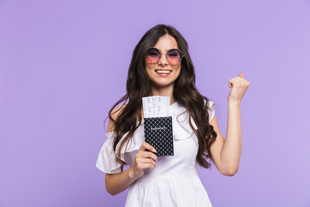 夏の衣装とサングラスを身に着けている美しい興奮した若い女性は、紫色の壁の上に孤立して立って、航空券とパスポートを示しています