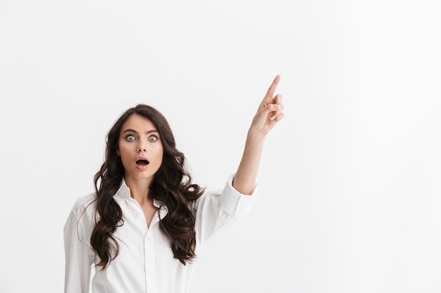 Beautiful excited young woman with long curly brunette hair wearing white shirt standing isolated over white wall, pointing finger at copy space