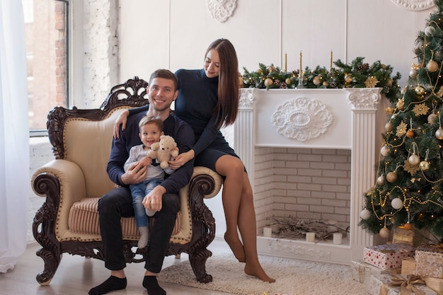 Beautiful family mom, dad and baby celebrate christmas by the new year tree with gifts and garland next to the fireplace.