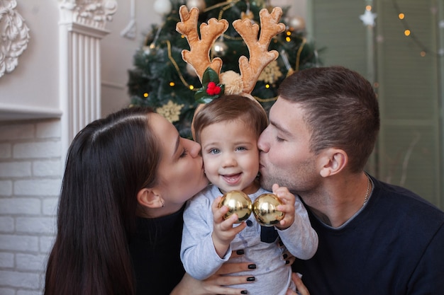 Bella mamma e papà di famiglia ridono e baciano il bambino con le corna di cervo sullo sfondo dell'albero di natale.