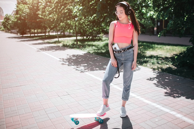 Beautiful and fantastic girl is standing on street in a sunny day and posing. She is holding one foot on skate. There is a music player in her pnats and heapsonhes on her head.