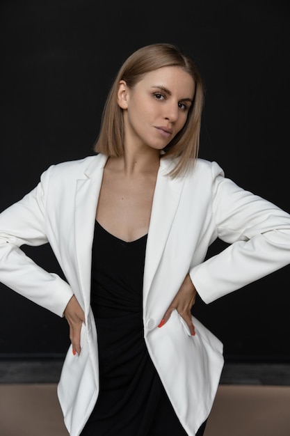 Beautiful fashion caucasian woman in a black dress and white jacket in the studio on a black background looking at the camera