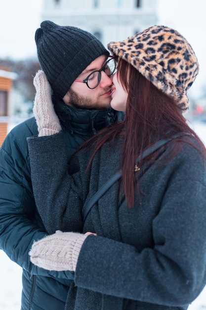写真 ジャケットと帽子を着て雪が降る街で抱きしめてキスするファッショナブルな冬の服を着た美しいファッションカップル