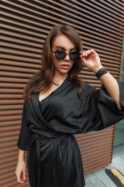 Beautiful fashion fresh brunette woman with hairstyle in a fashionable elegant black dress puts on cool sunglasses and walks in the city near the metal wall