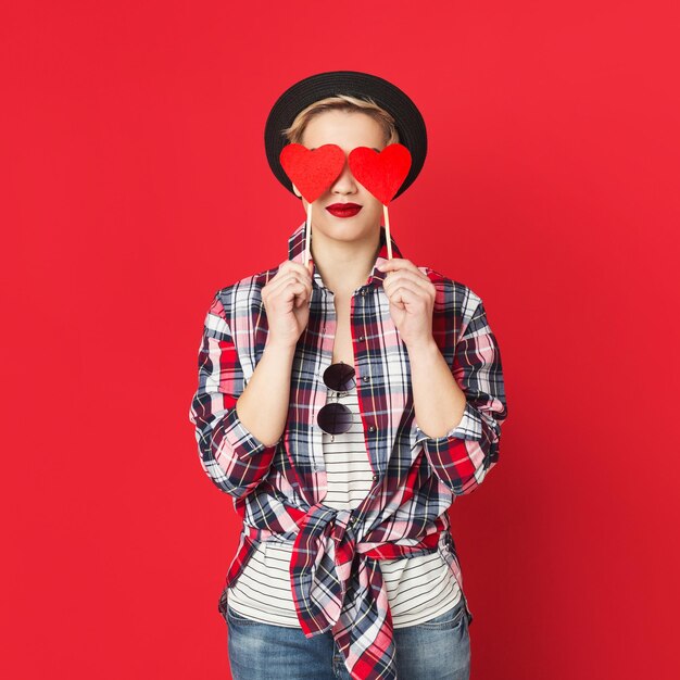 Beautiful fashion girl holding red paper hearts covering her eyes, red studio background