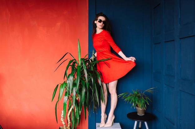 Beautiful fashion girl with long hair,  in sunglasses and red elegant dress posing on blue red wall between green trees.