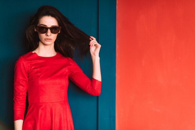 Beautiful fashion girl with long hair in sunglasses and red elegant dress posing on blue red wall.