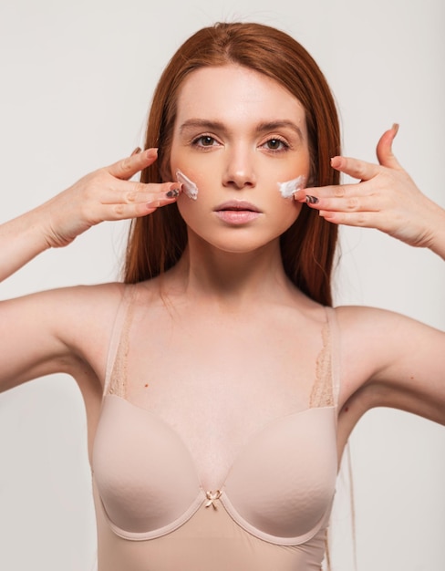 Beautiful fashion model woman with red hair and fresh clean skin in a beige bra applies a cream on her face in the studio Beauty and skin care