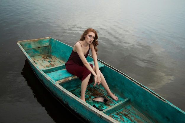 Beautiful fashion redhead woman sit in boat. Beauty romantic portrait girl in red vinous dress in wooden boat on lake