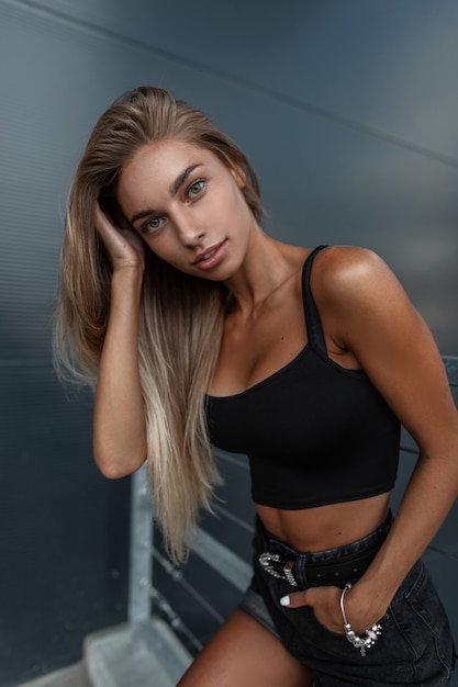 Beautiful fashion sexual woman model with a pretty face in denim shorts and a black T-shirt with a female jewelry bracelet  on hand on the street near metal building