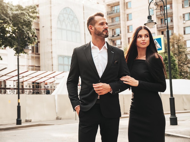 Beautiful fashion woman and her handsome elegant boyfriend in suit Sexy brunette model in black evening dress Fashionable couple posing in the street in Europe Brutal man and his female outdoors