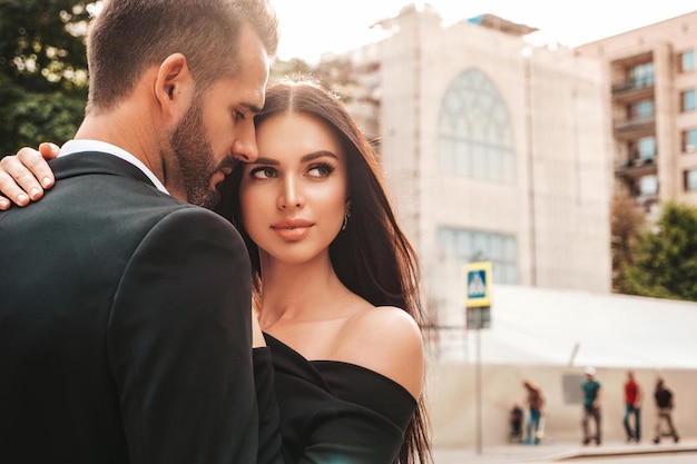 Beautiful fashion woman and her handsome elegant boyfriend in suit Sexy brunette model in black evening dress Fashionable couple posing in the street in Europe Brutal man and his female outdoors