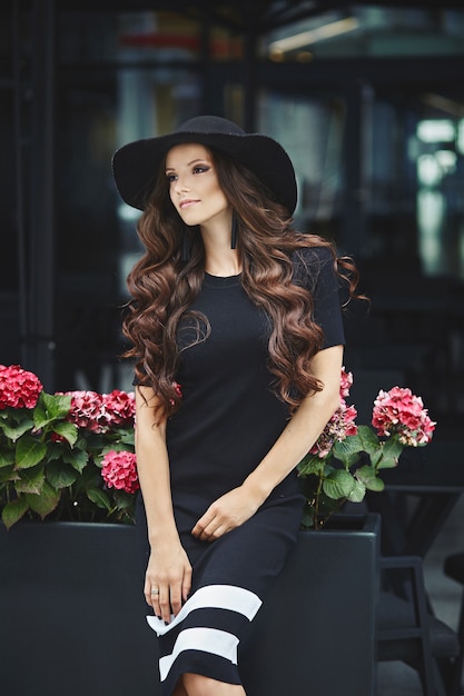 Beautiful, fashionable brunette model girl in black dress and hat posing outdoors, spring