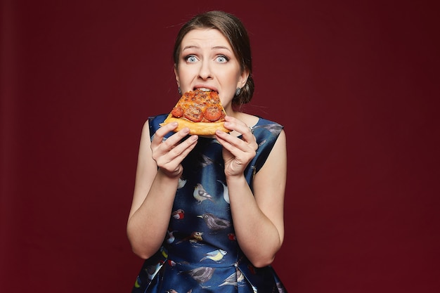 Beautiful and fashionable brunette model girl in blue dress eating pizza isolated at vinous backgrou...