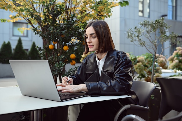 Beautiful fashionable brunette woman, copywriter, developer, freelancer, typing text on laptop, sitting at outdoor cafe with decorative tangerine trees on the background, enjoying distant online work