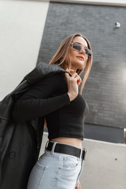 Красивая модная девушка в модной одежде с крутыми золотыми очками в черной футболке с джинсами и пальто, идущими по улице
